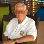 Disney Legend Bob Gurr smiles while seated in an outdoor wicker chair, wearing a white polo shirt with a Disneyland 50th anniversary crest.