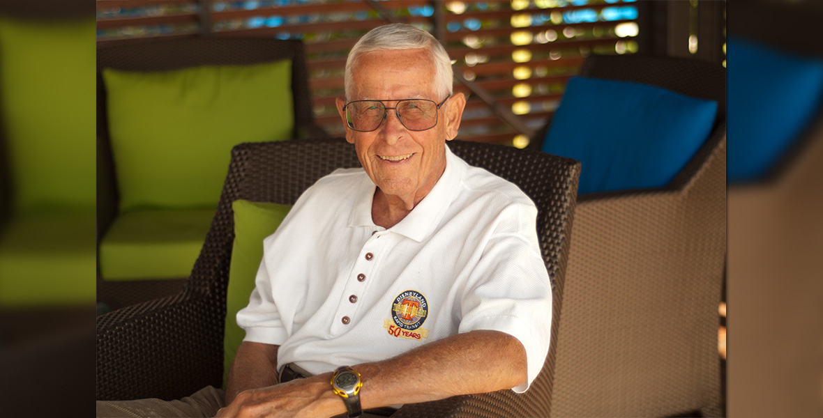Disney Legend Bob Gurr smiles while seated in an outdoor wicker chair, wearing a white polo shirt with a Disneyland 50th anniversary crest.