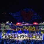 The Disney Destiny lit up at night during its christening ceremony, with a drone show overhead and performers on a stage in front of the ship.