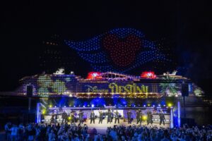 The Disney Destiny lit up at night during its christening ceremony, with a drone show overhead and performers on a stage in front of the ship.