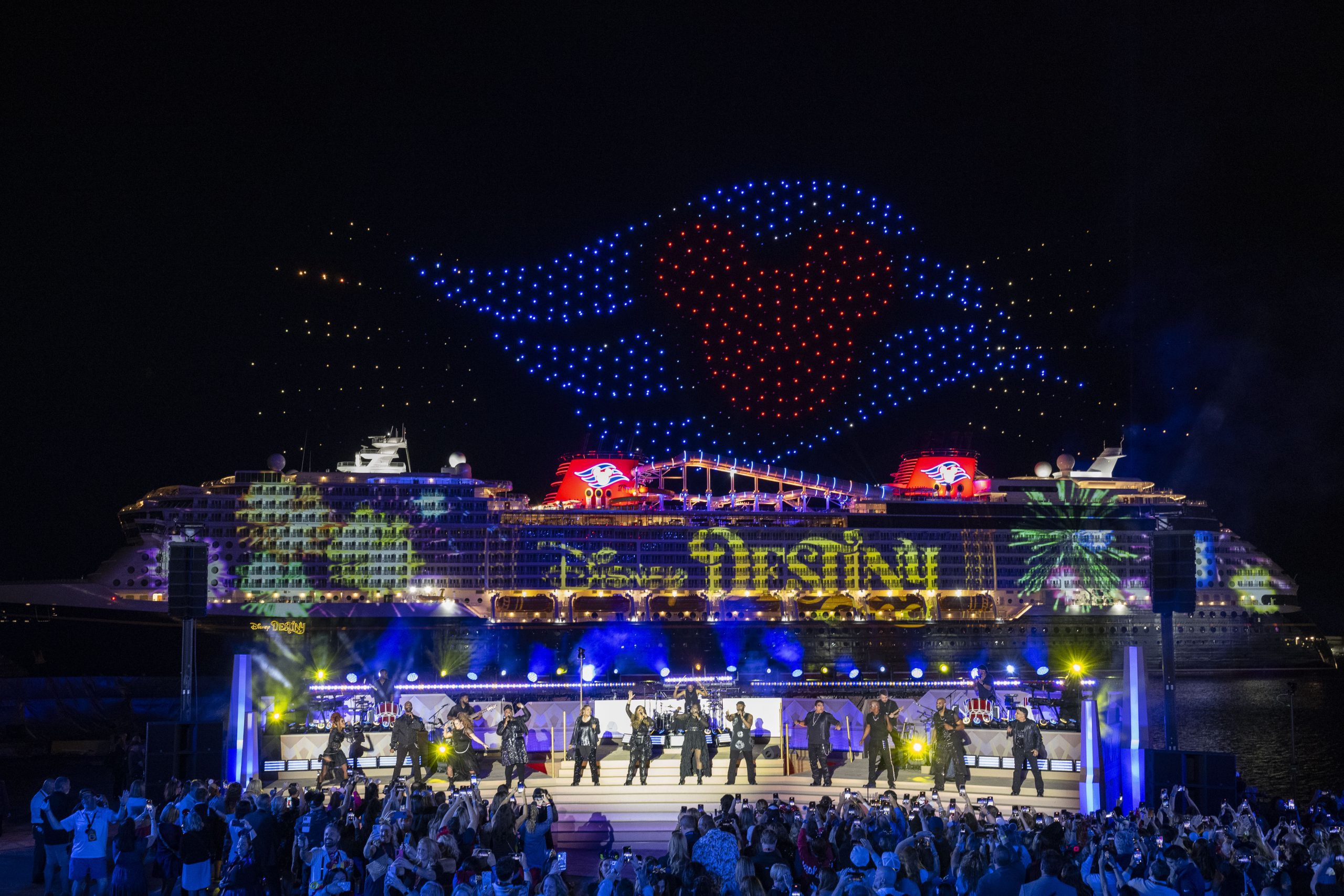 The Disney Destiny lit up at night during its christening ceremony, with a drone show overhead and performers on a stage in front of the ship.