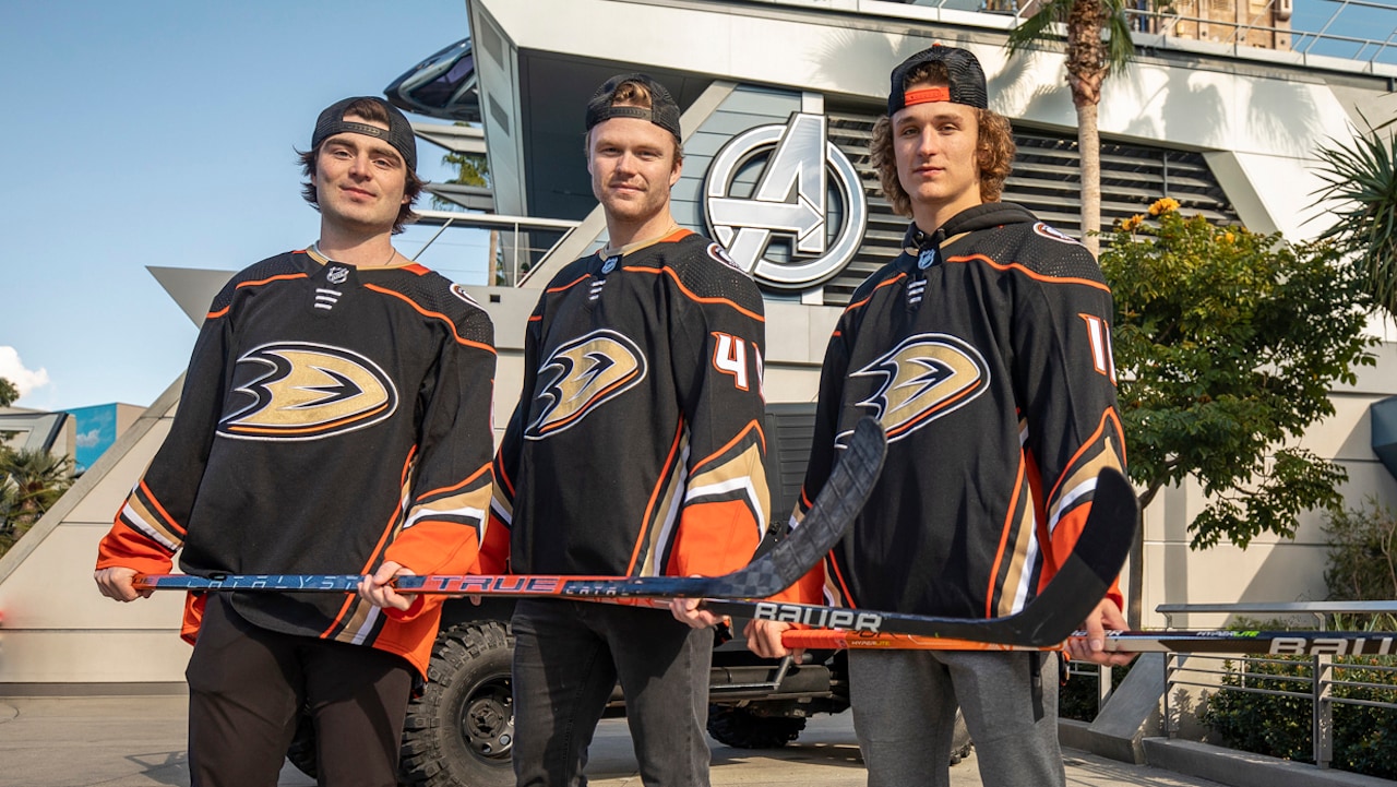 Three Anaheim Ducks players in team jerseys hold hockey sticks outside Avengers Campus at Disney California Adventure