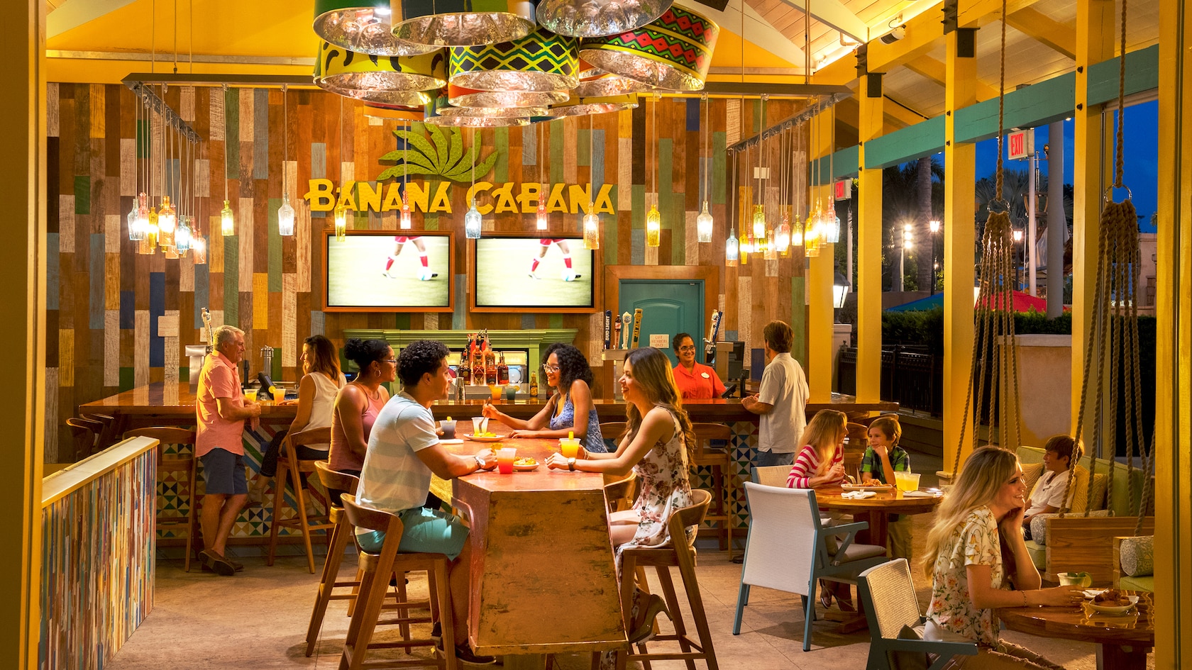 A bar with 2 TVs displayed on the wall, a sign that says Banana Cabana, a communal table and other tables with Guests sitting and eating