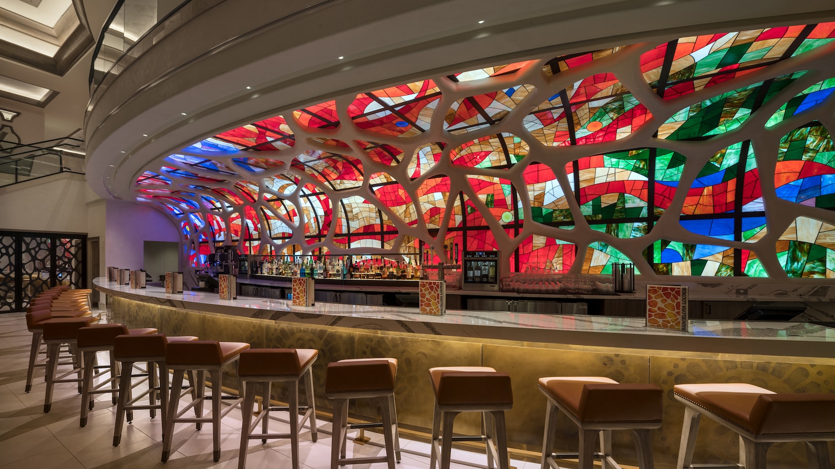 Stools in front of a long bar with a brilliant stained glass wall boasting many colors behind the bar