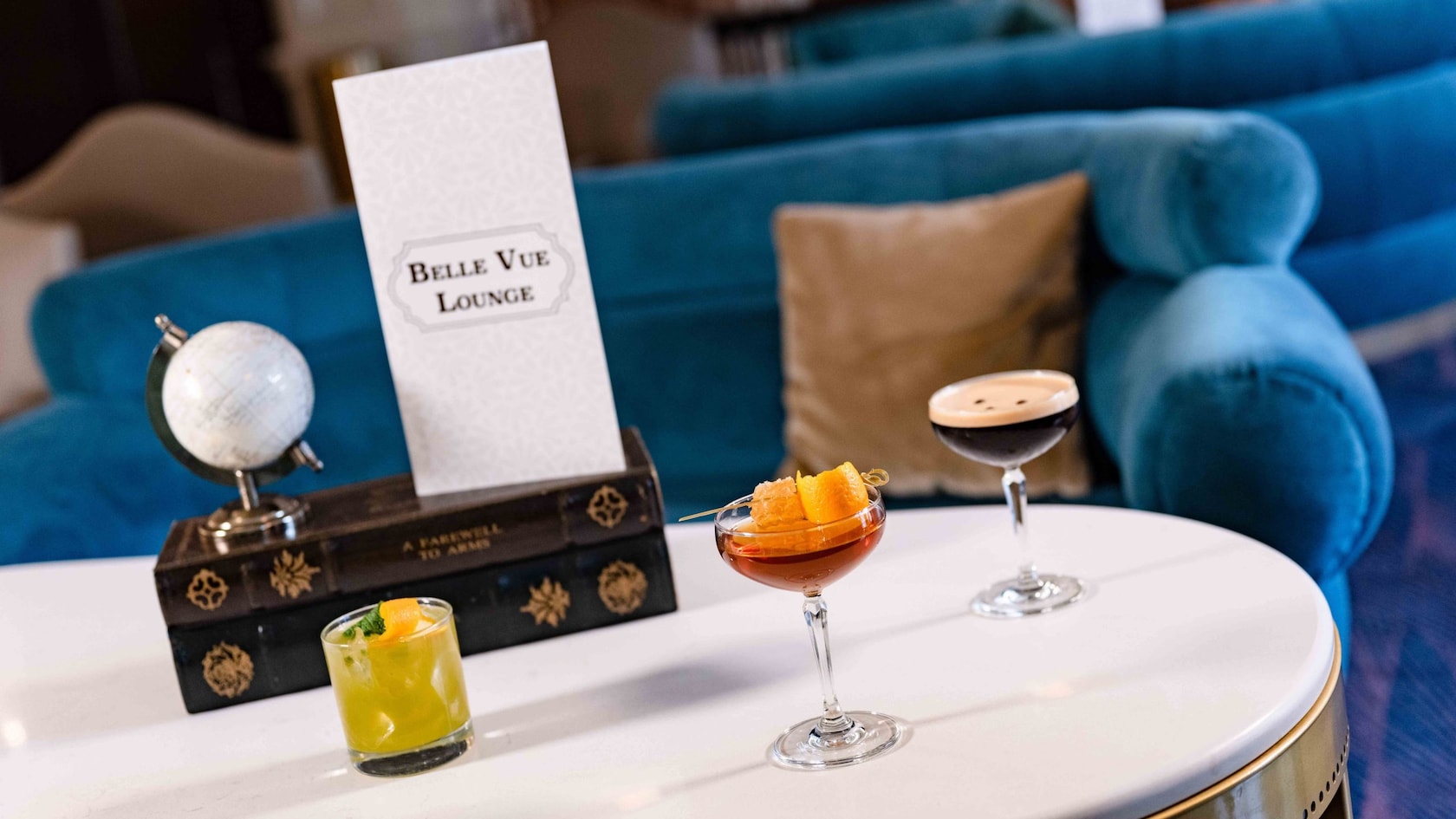 A table featuring 3 cocktails and a menu that reads ‘Belle Vue Lounge’ across the front at Disney’s Boardwalk Inn