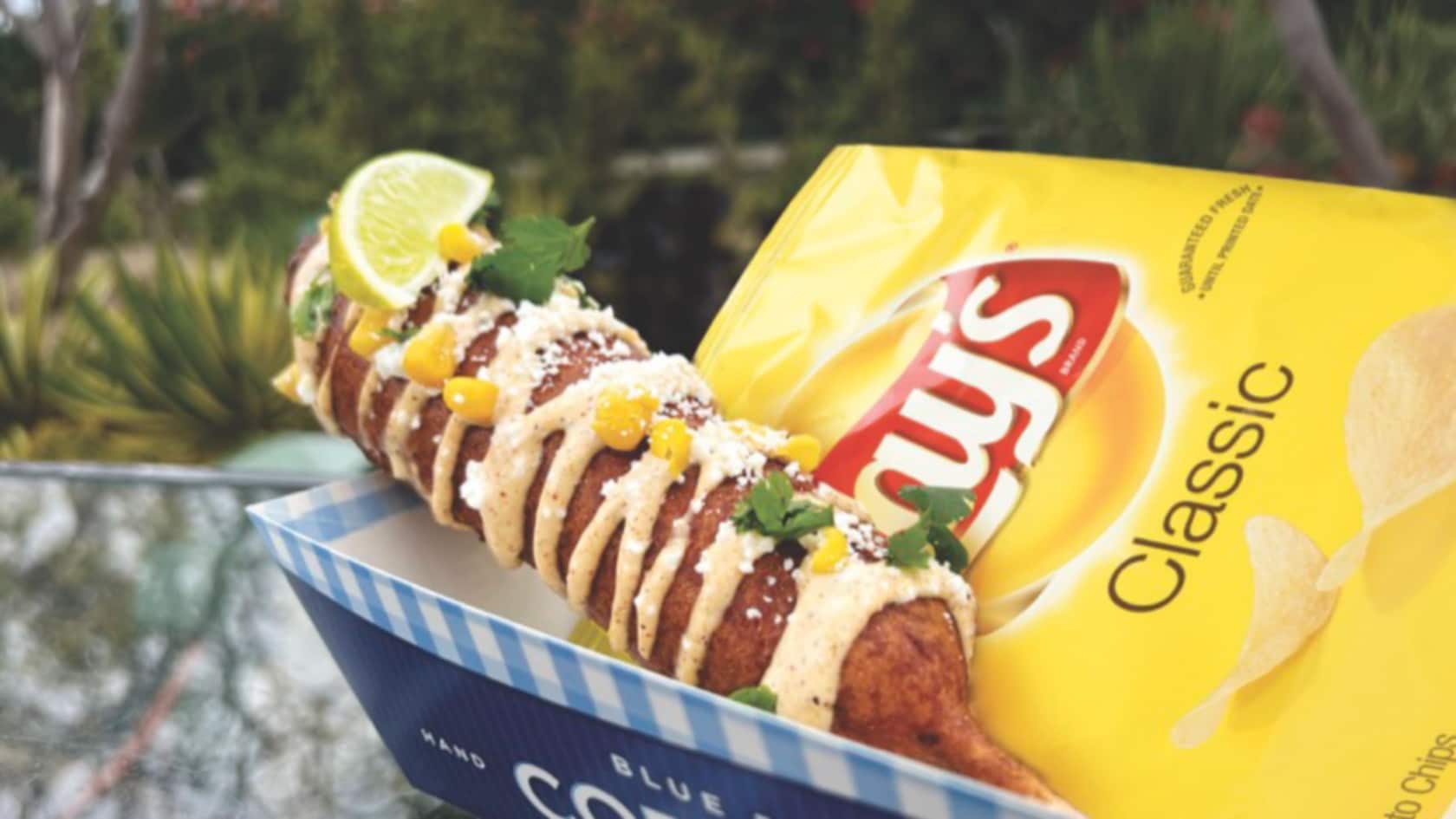A Mexican Street Dog featuring garlic mayo and corn alongside a bag of potato chips