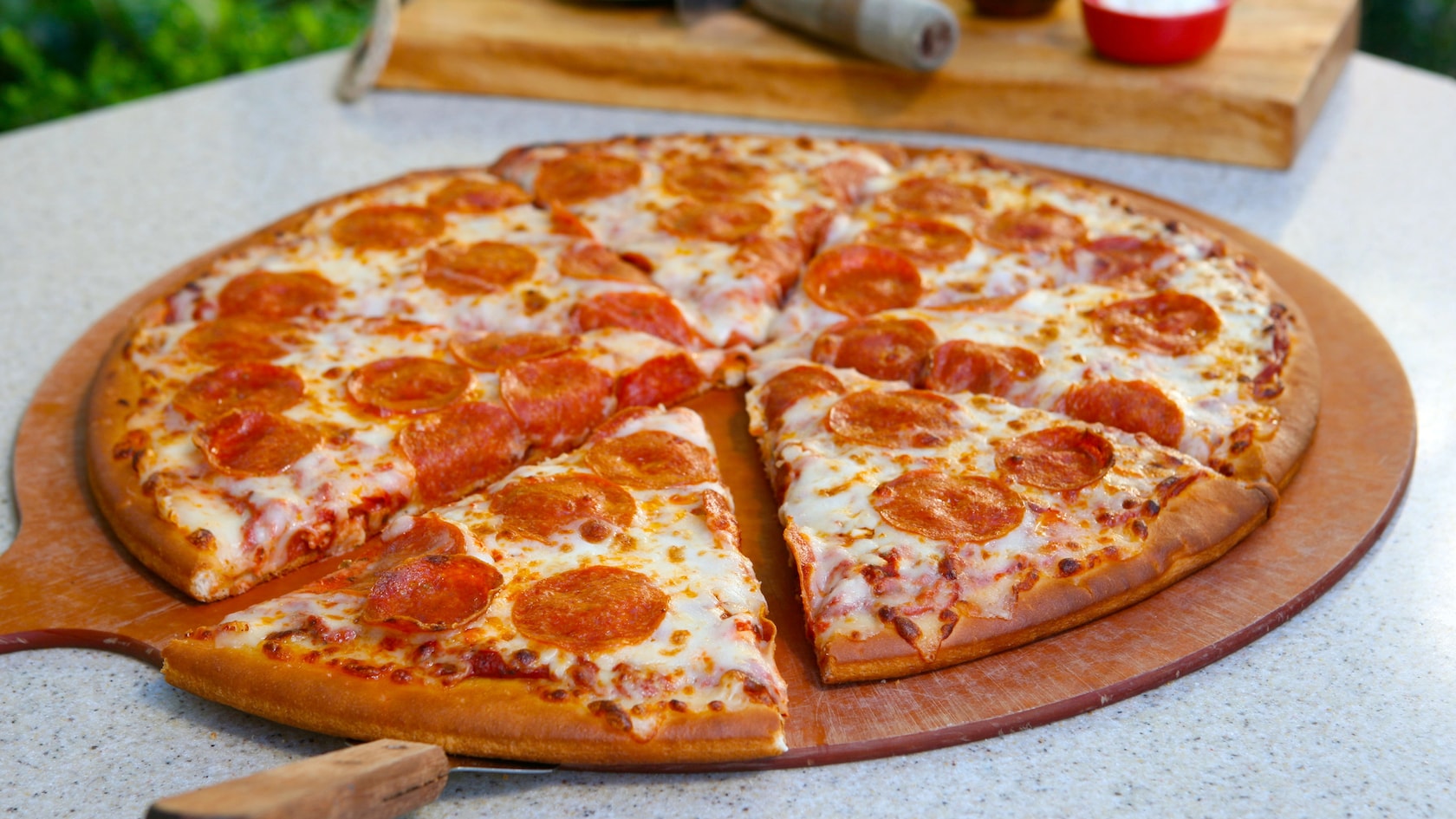 A fresh out of the oven, sliced pepperoni pizza on a wooden pizza board sits on a table outdoors