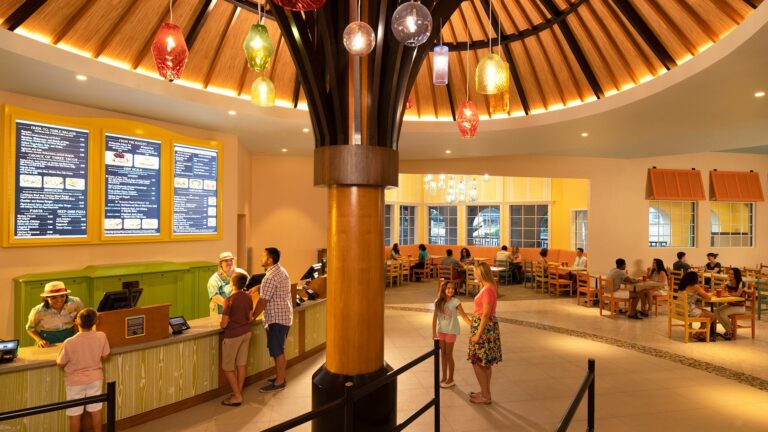 A cafeteria with a large umbrella sculpture, hanging lights, a large menu displayed on the wall, Guests ordering at the front registers and other Guests sitting and eating