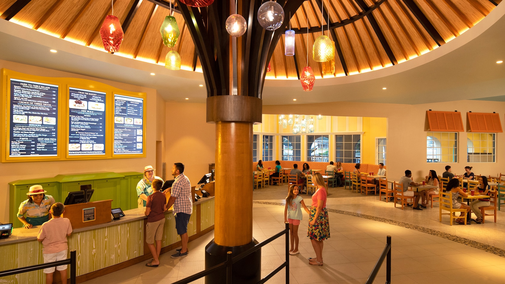 A cafeteria with a large umbrella sculpture, hanging lights, a large menu displayed on the wall, Guests ordering at the front registers and other Guests sitting and eating