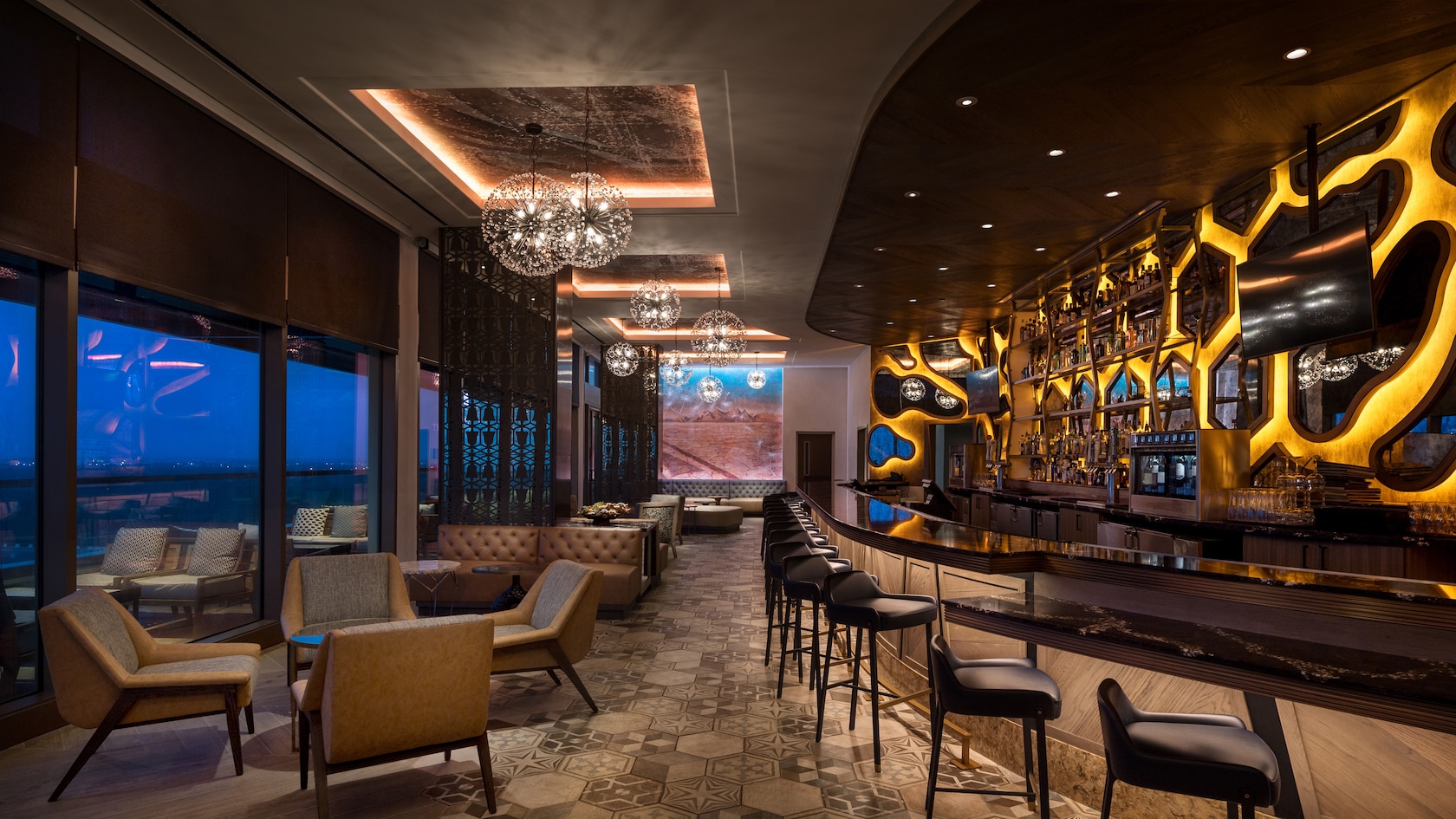 Wooden tables and upholstered chairs line against a wall of windows opposite a long bar with tall stools in a carpeted room with a recessed ceiling and contemporary chandeliers and dramatic lighting