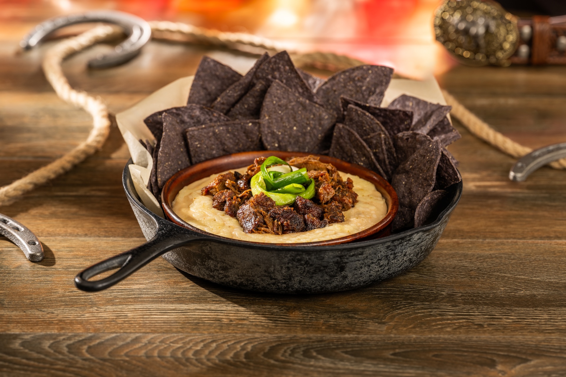 Queso fundido topped with burnt ends and sliced jalapeño in a skillet with blue corn tortilla chips