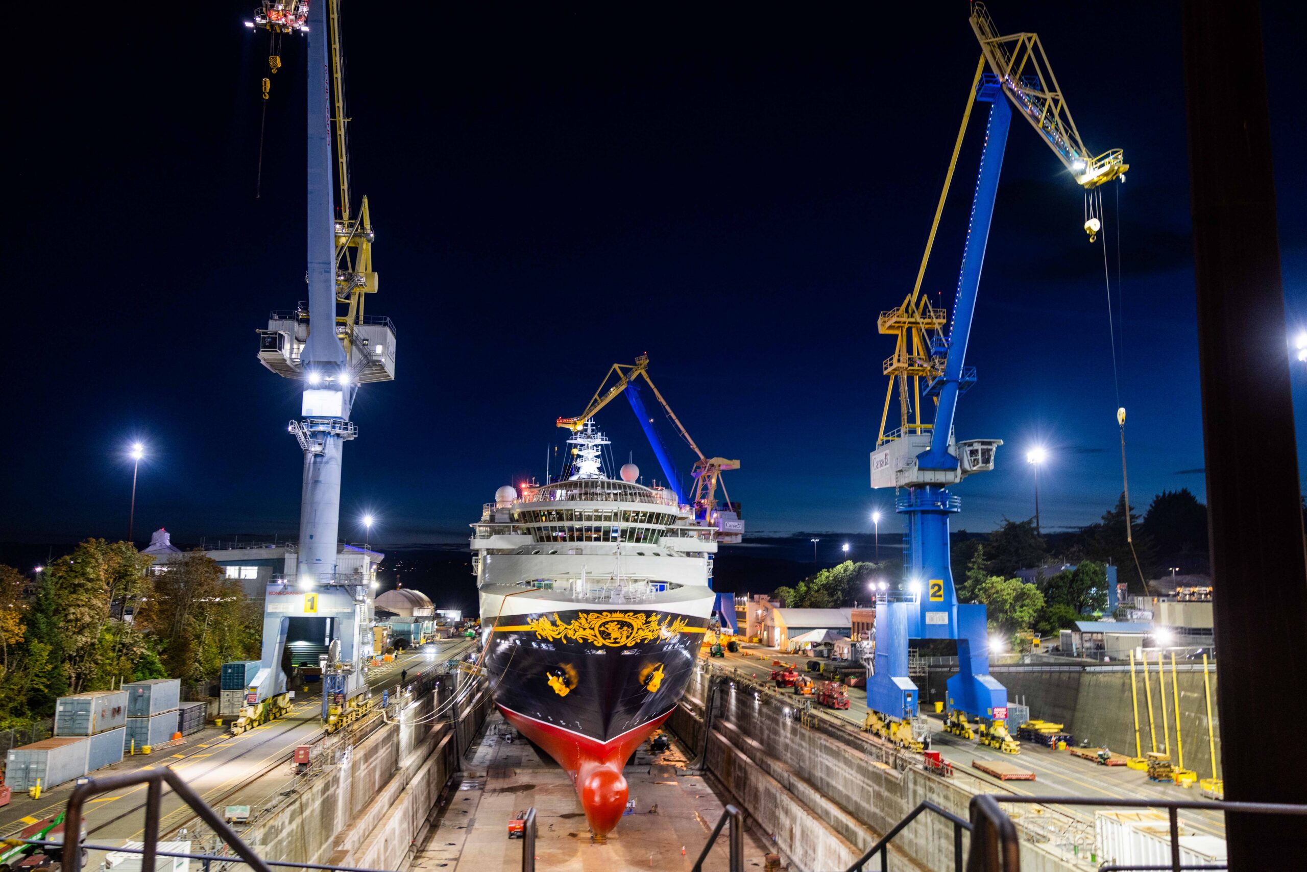 A Disney Cruise Line ship in dry dock at night with cranes on either side