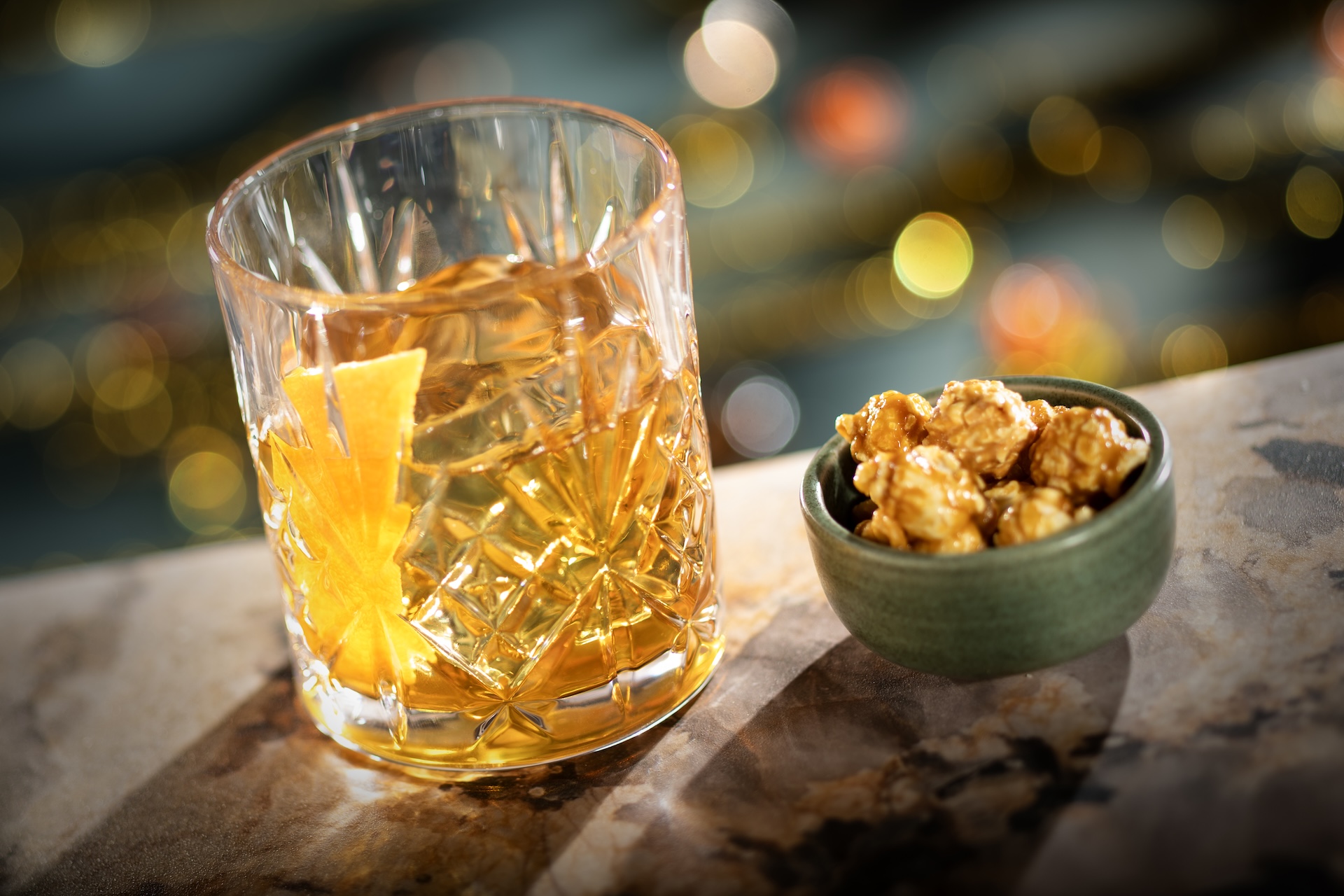 A Hurly-Burly cocktail in a rocks glass with an orange twist served beside a bowl of caramel popcorn