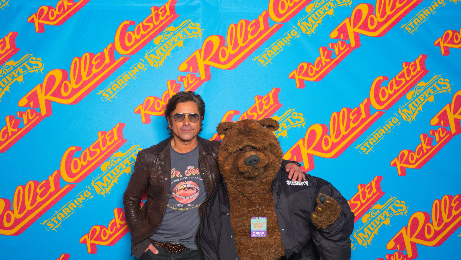 John Stamos posing with Fozzie Bear in a security jacket on the Rock 'n' Roller Coaster Starring The Muppets step and repeat at Disney's Hollywood Studios