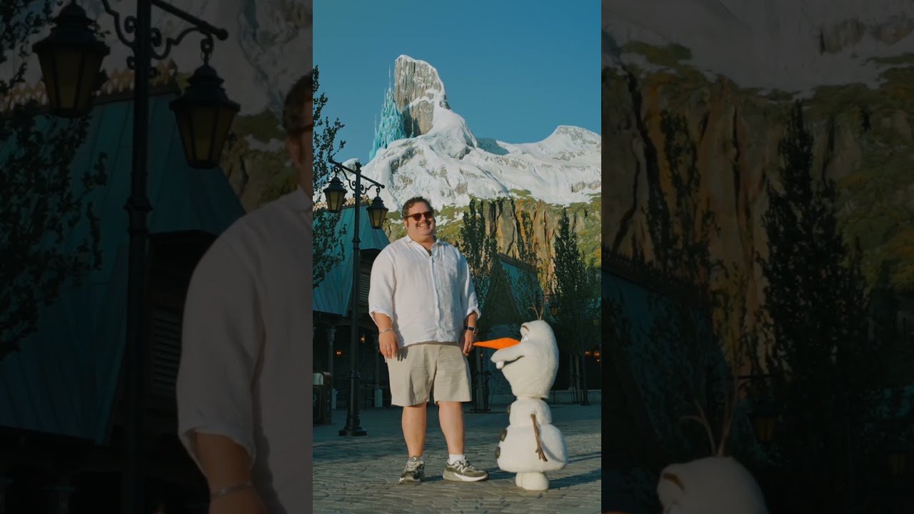 Josh Gad smiles next to the Olaf animatronic in World of Frozen at Disney Adventure World, with North Mountain behind them
