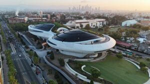 Aerial view of the Lucas Museum of Narrative Art building in Exposition Park, with the downtown Los Angeles skyline behind it