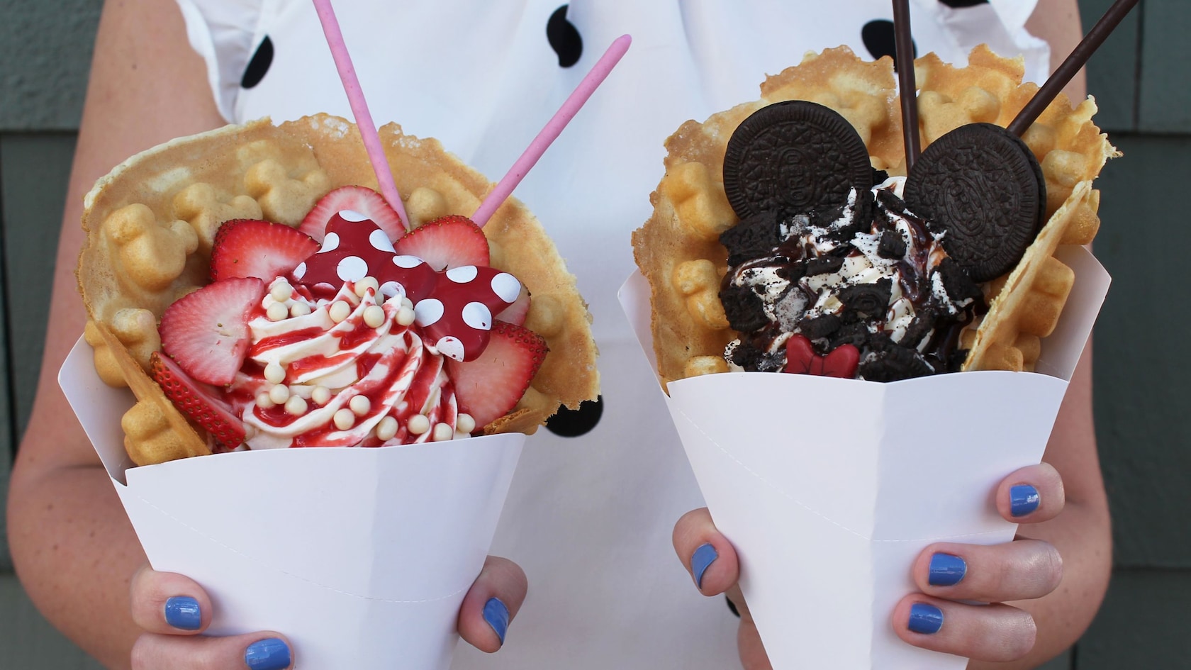 A woman holding a waffle sundae in each hand, one sundae topped with strawberries and the other topped with cookies