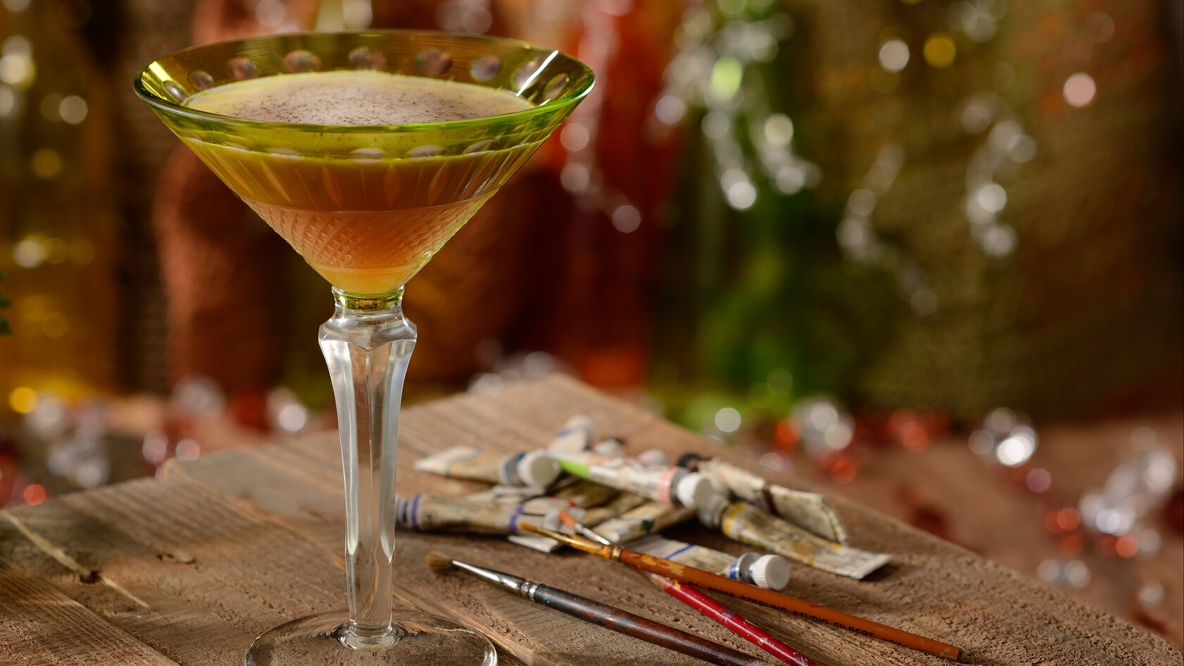 An elegant martini glass filled with a Night Monkey cocktail on a table beside tubes of watercolor paints and brushes