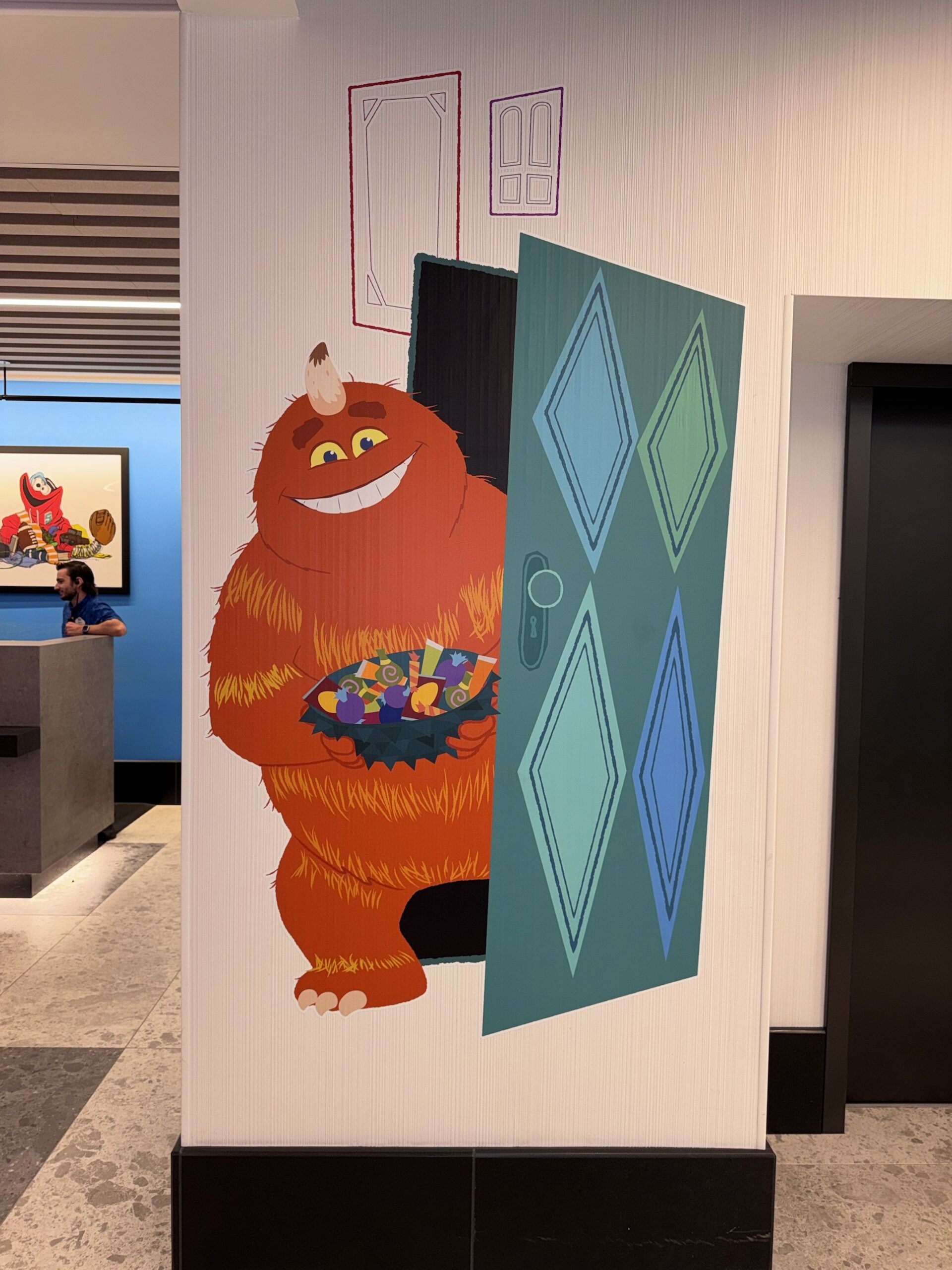 Wall artwork of a furry orange monster holding a bowl of candy beside an open door at Pixar Place Hotel