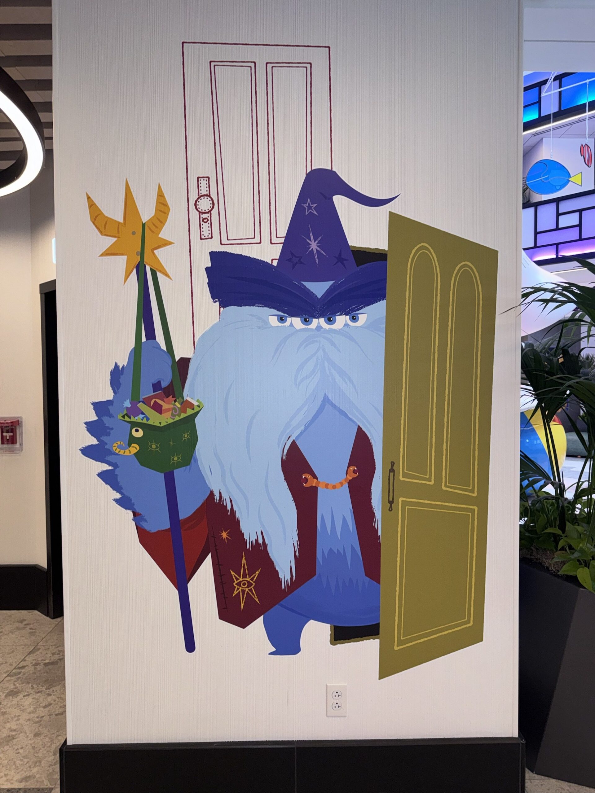 Wall artwork of a four-eyed blue monster dressed as a wizard with a staff and candy bag stepping out of a door at Pixar Place Hotel