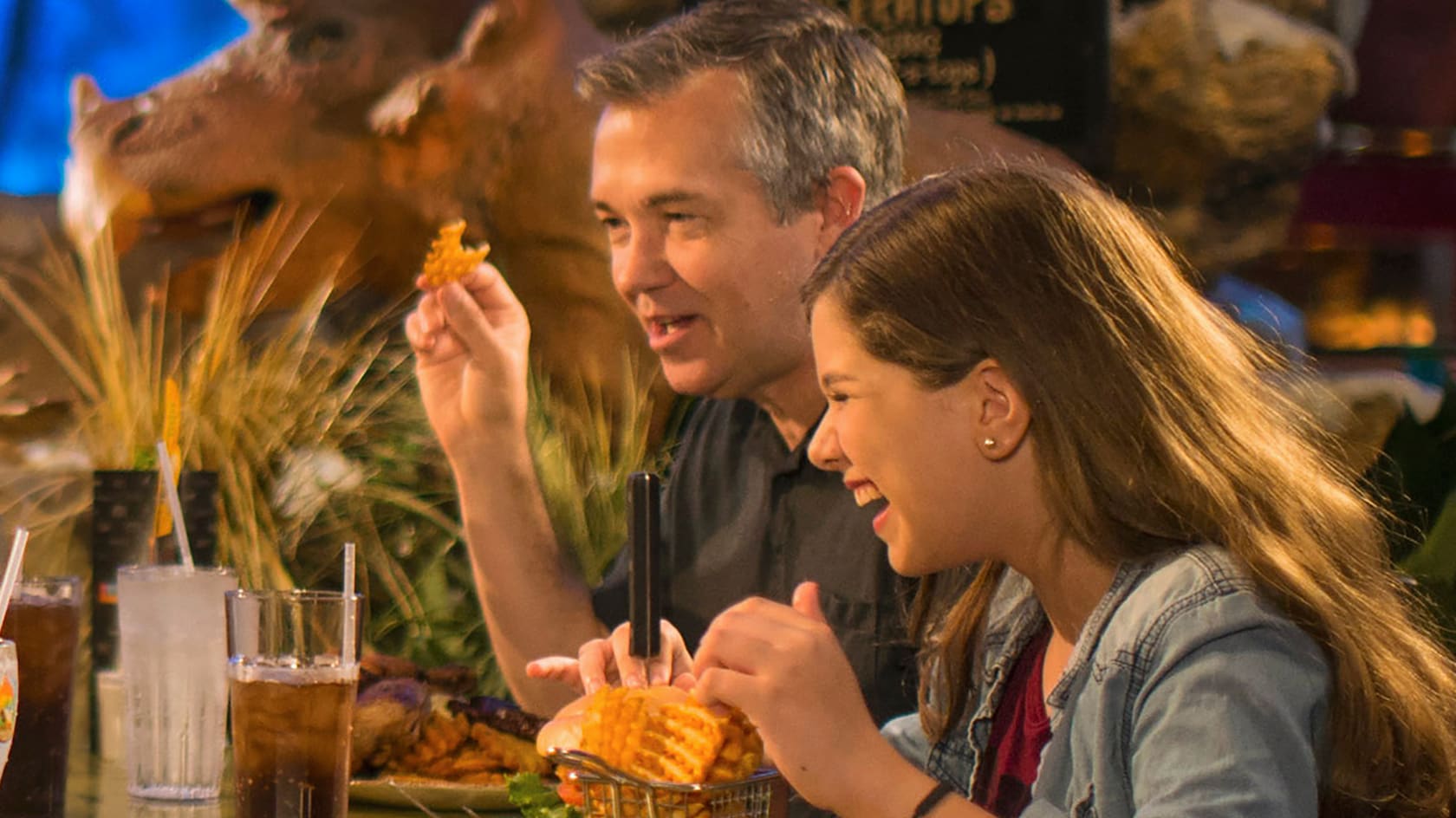 A girl and her dad enjoy waffle fries seated at their table with Dinosaur themed decor in the background