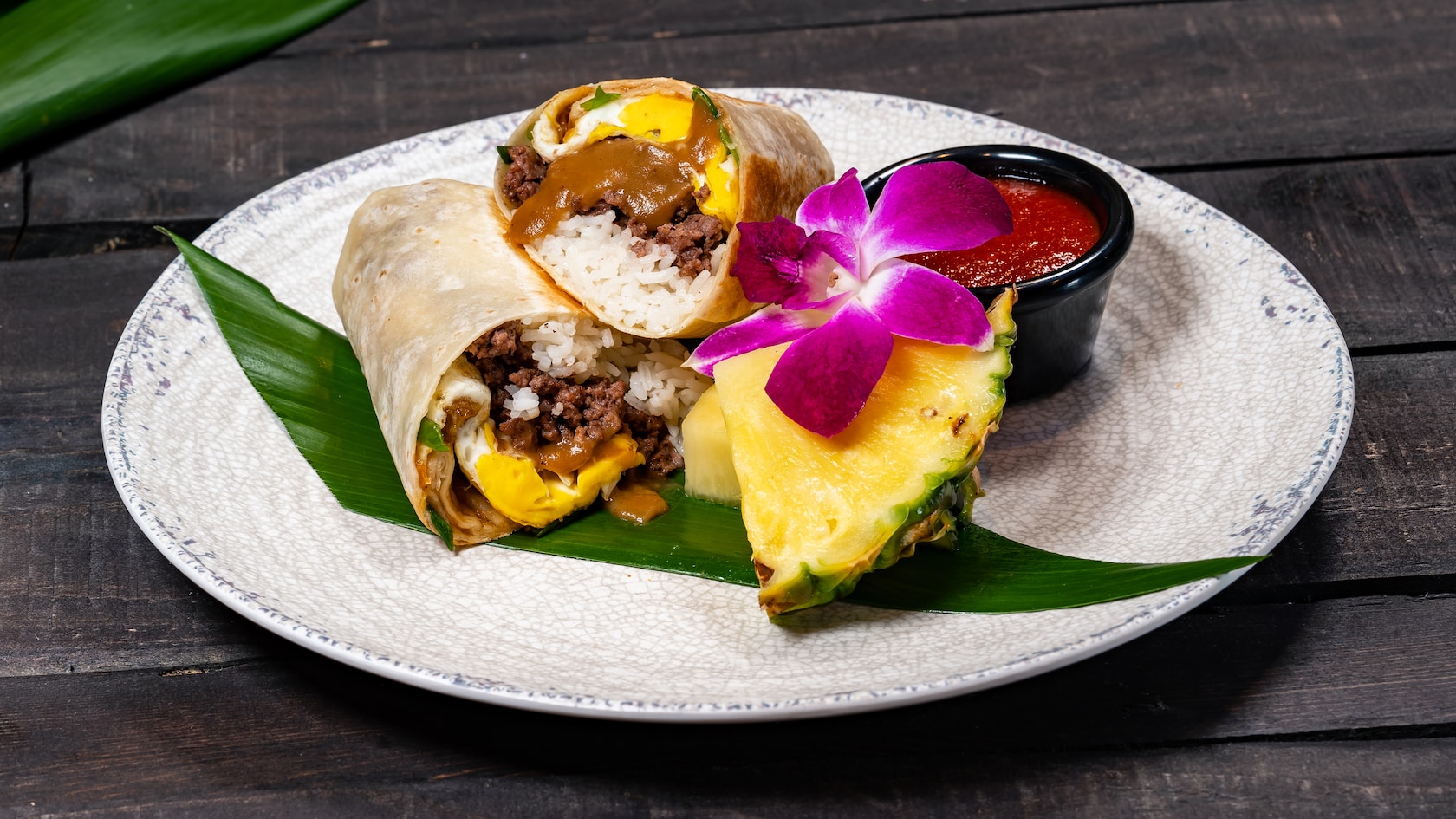 A burrito filled with rice, ground beef, fried egg and gravy on a plate with a cup of sauce, pineapple wedges and an orchid flower