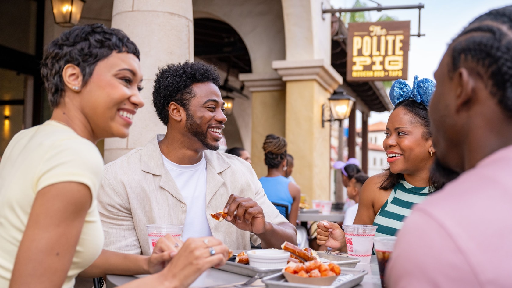 Four Guests enjoying a meal together at a table outdoors under a sign with the words ‘The Polite Pig’