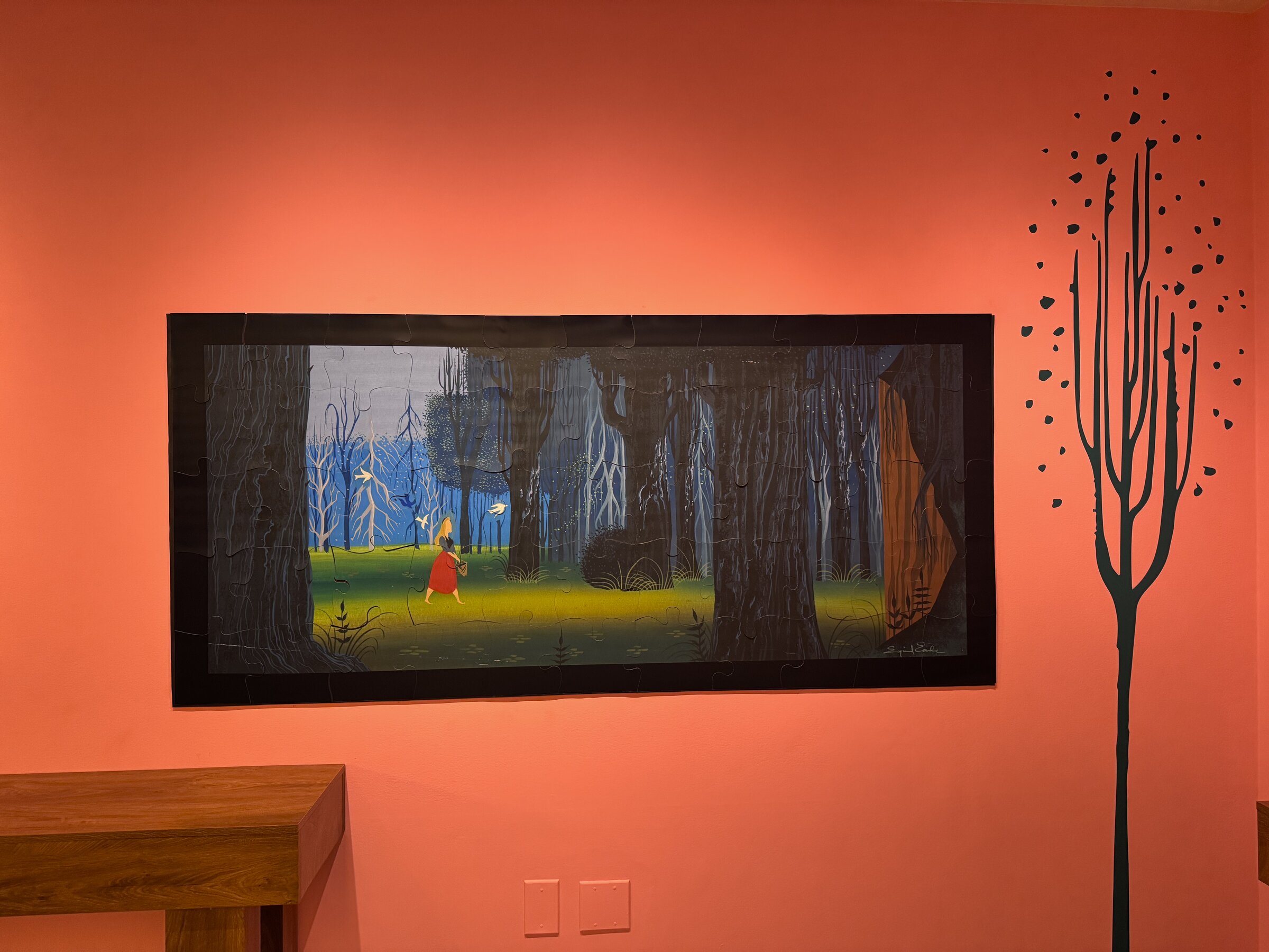 A wall-size magnetic puzzle of Eyvind Earle's Sleeping Beauty concept art showing a small figure in red in a blue forest, with pieces missing