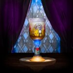 The Be Our Guest Goblet, with a stained-glass style image of Belle and the Beast dancing on the bowl, a lid and straw, a glowing blue section in the stem above a red rose under a clear dome, and a gold base