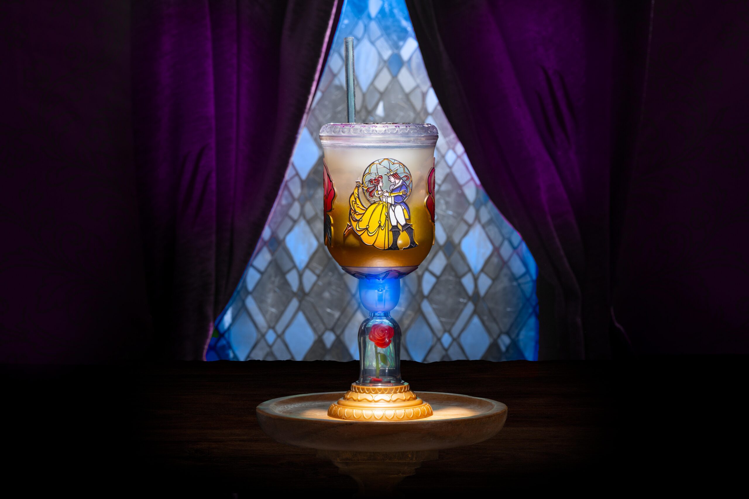 The Be Our Guest Goblet, with a stained-glass style image of Belle and the Beast dancing on the bowl, a lid and straw, a glowing blue section in the stem above a red rose under a clear dome, and a gold base