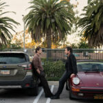 Pedro Pascal and a co-star lean against parked cars under palm trees in a still from Searchlight Pictures' Behemoth!