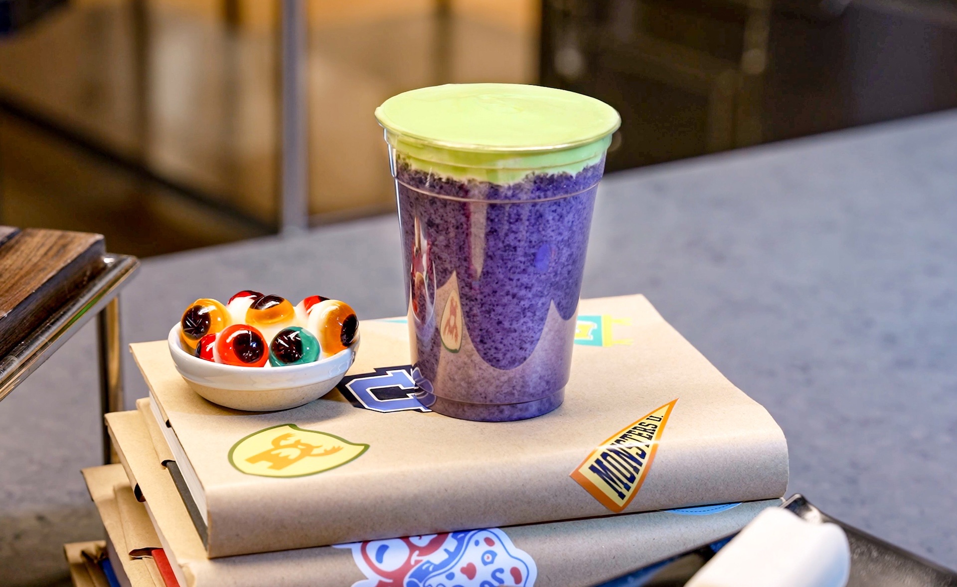A purple frozen drink with a green foam topping on a stack of books covered in Monsters University stickers, next to a small bowl of eyeball candies. (Disney)