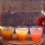 Bountiful Valley Cocktail Flight, three cocktails on a round wooden board, one garnished with a peach gummy ring and one with a strawberry