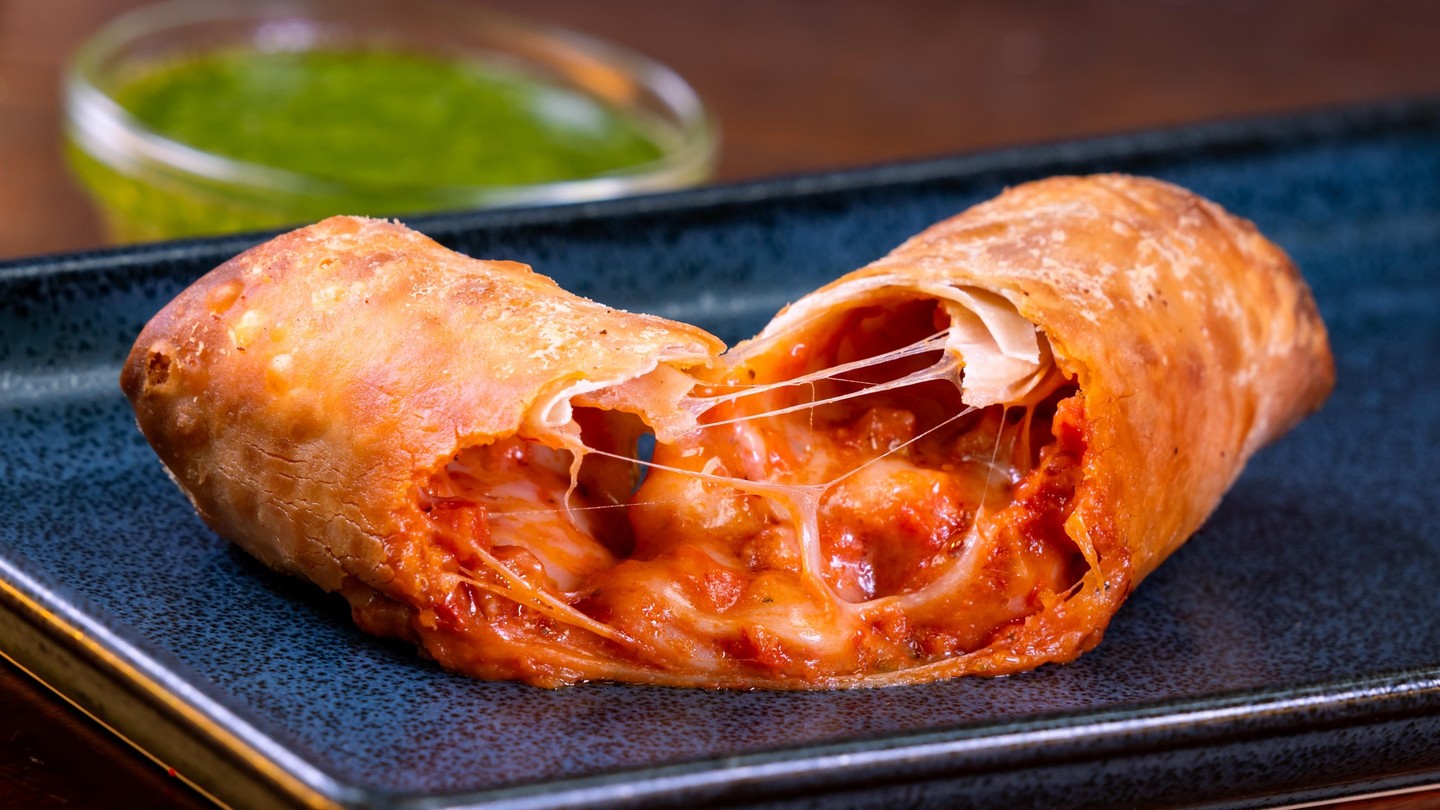 Pepperoni Pizza Egg Roll cut in half with melted cheese stretching between the pieces, with a cup of pesto dipping sauce behind it