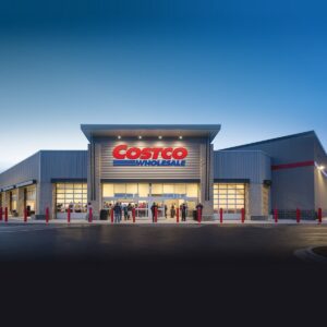 Exterior of a Costco Wholesale warehouse at dusk, with shoppers gathered at the lit entrance