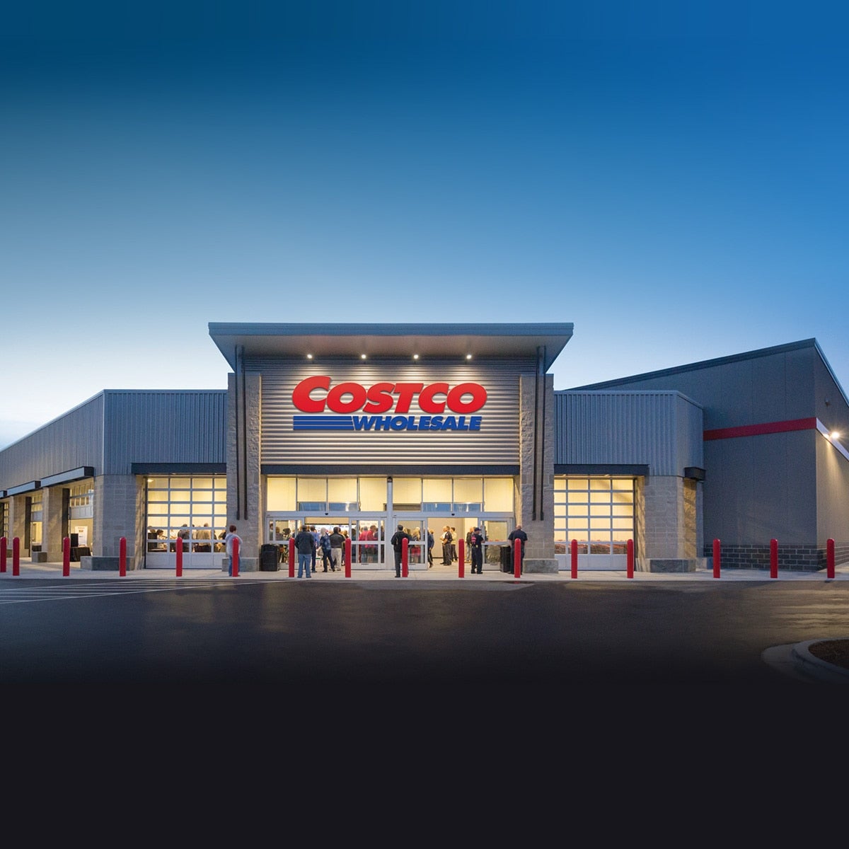 Exterior of a Costco Wholesale warehouse at dusk, with shoppers gathered at the lit entrance