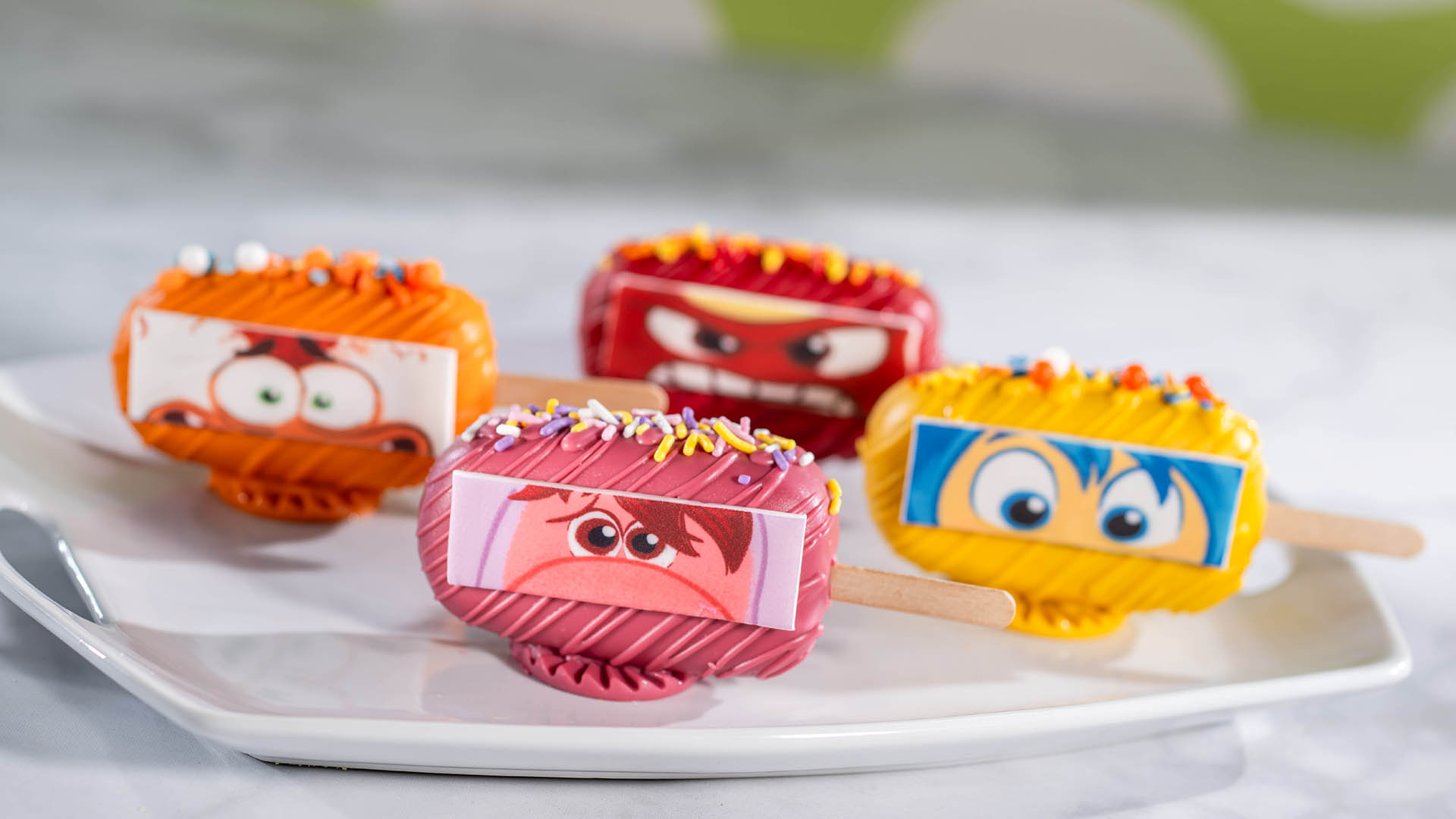 Four Inside Out cake popsicles in orange, red, pink and yellow, each decorated with an emotion's eyes