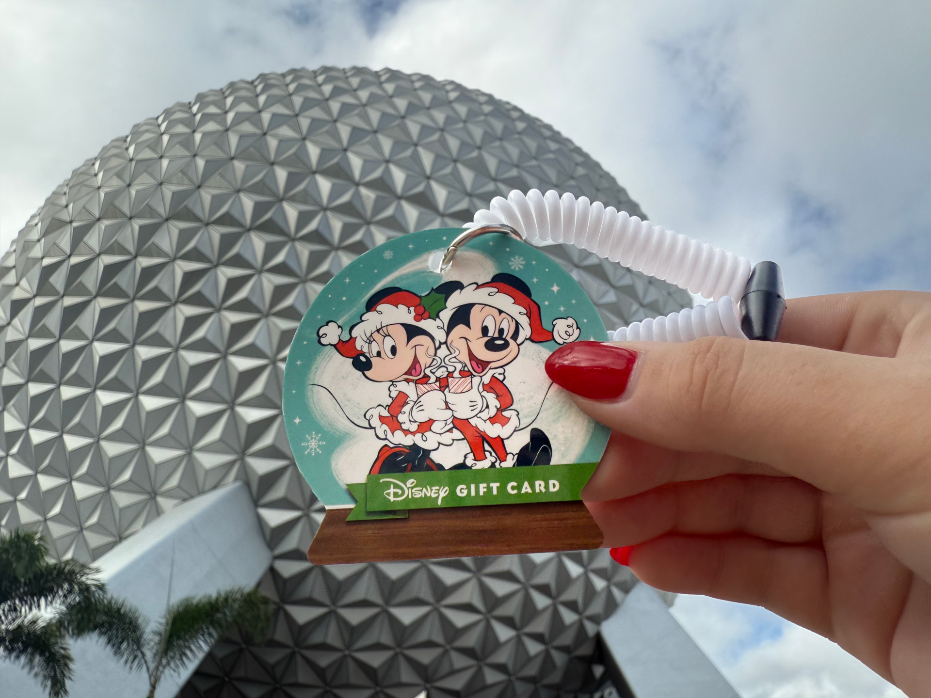 A Mickey and Minnie Mouse holiday Disney Gift Card held up in front of Spaceship Earth at EPCOT