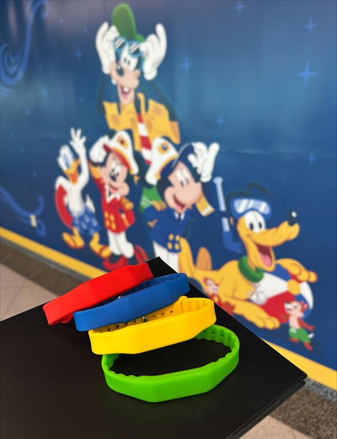 Red, blue, yellow and green Key to the World wristbands in front of a Disney Cruise Line mural of Mickey, Minnie, Goofy, Donald and Pluto