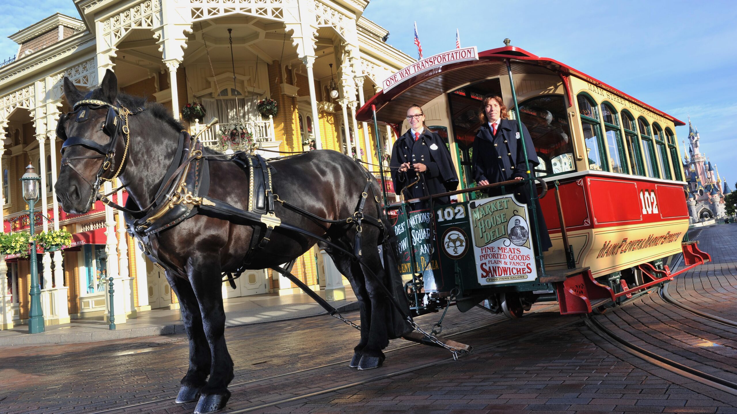 A dark horse in harness stands with Horse-Drawn Streetcar No. 102 and two cast members on Main Street, U.S.A. at Disneyland Paris