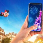 A hand holding a phone showing Sleeping Beauty Castle and fireworks in front of the Eiffel Tower, with a Minnie Mouse balloon overhead