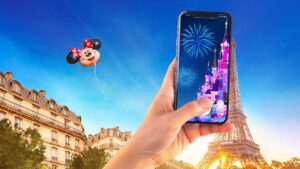 A hand holding a phone showing Sleeping Beauty Castle and fireworks in front of the Eiffel Tower, with a Minnie Mouse balloon overhead