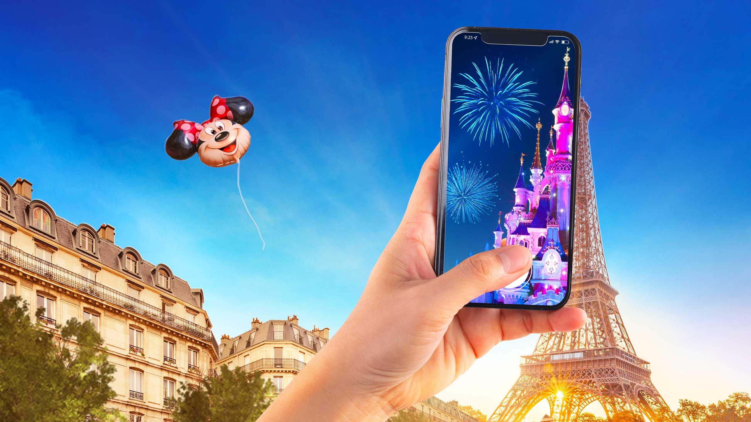 A hand holding a phone showing Sleeping Beauty Castle and fireworks in front of the Eiffel Tower, with a Minnie Mouse balloon overhead