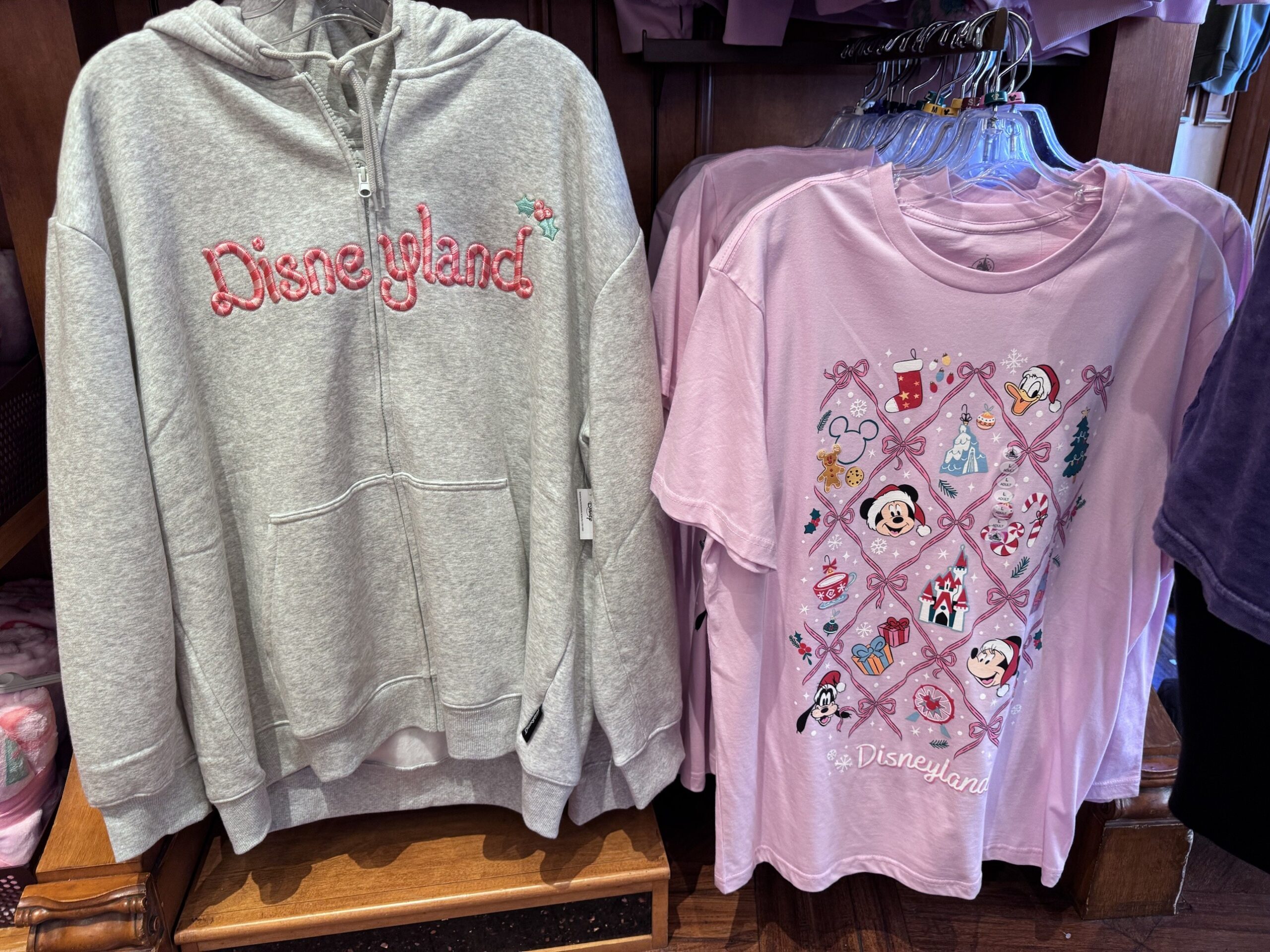 Gray zip hoodie with candy cane Disneyland embroidery next to a pink tee with Mickey, Minnie, Donald and Goofy in Santa hats inside a ribbon lattice
