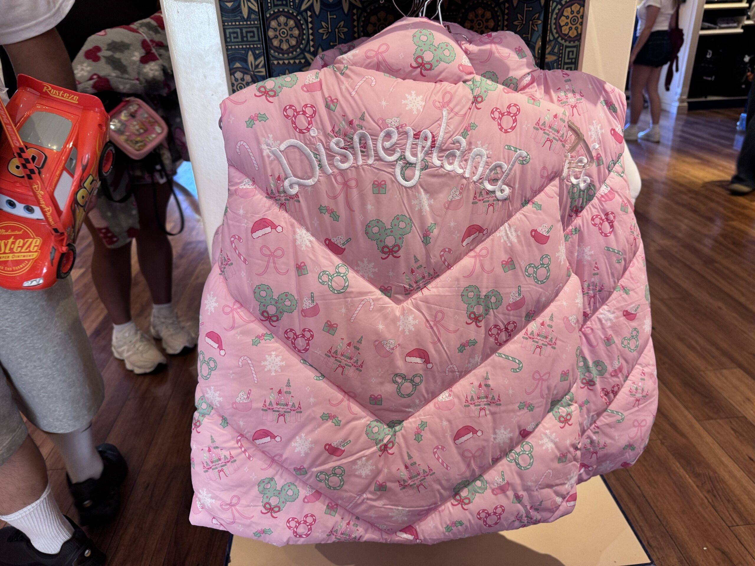 Back of a pink quilted puffer vest embroidered with Disneyland in white script over a print of castles, cocoa mugs, Santa hats and wreath Mickeys