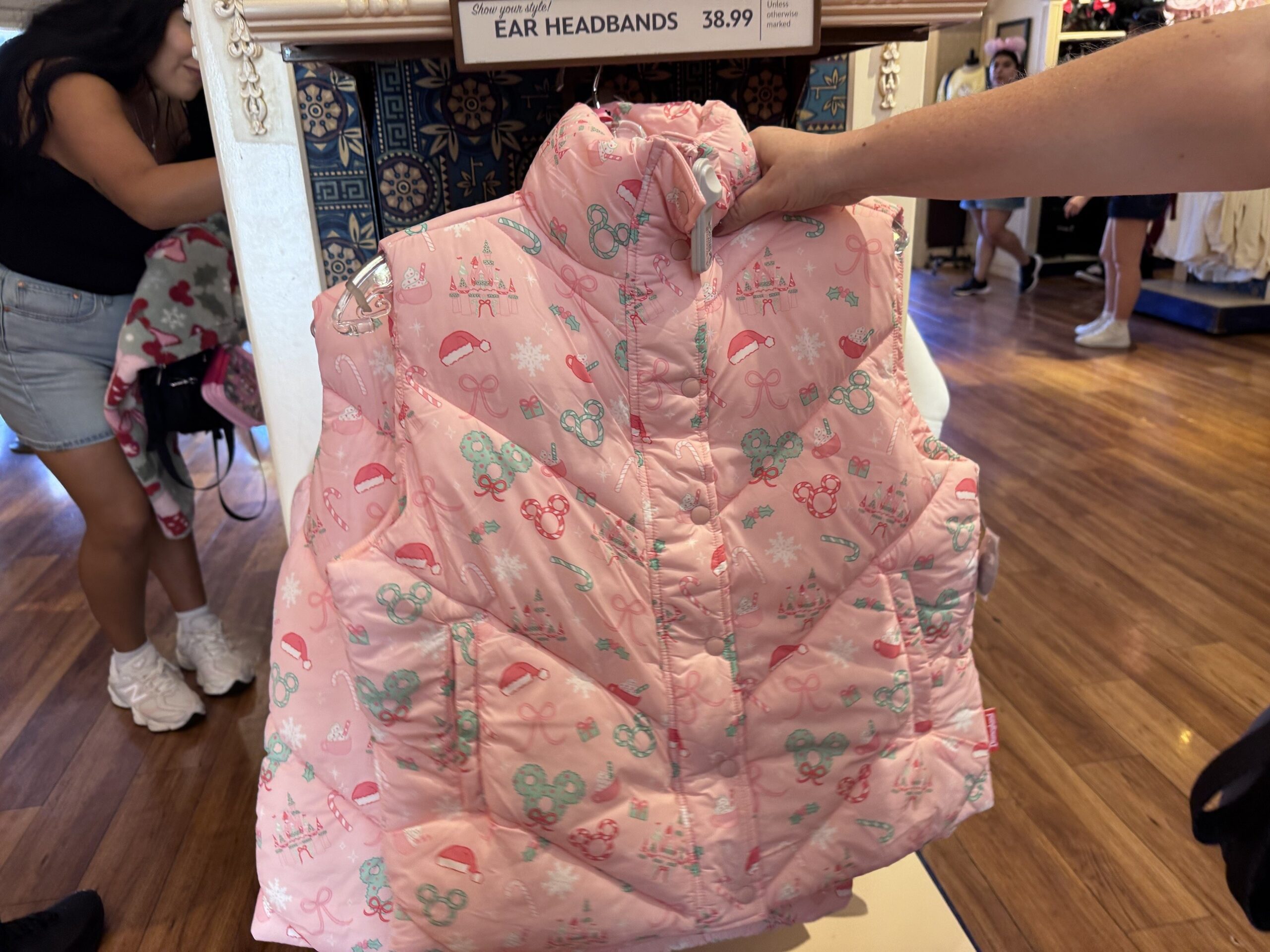 Front of a pink snap-front puffer vest with chevron quilting and a print of candy canes, wreath Mickeys, Santa hats and bows