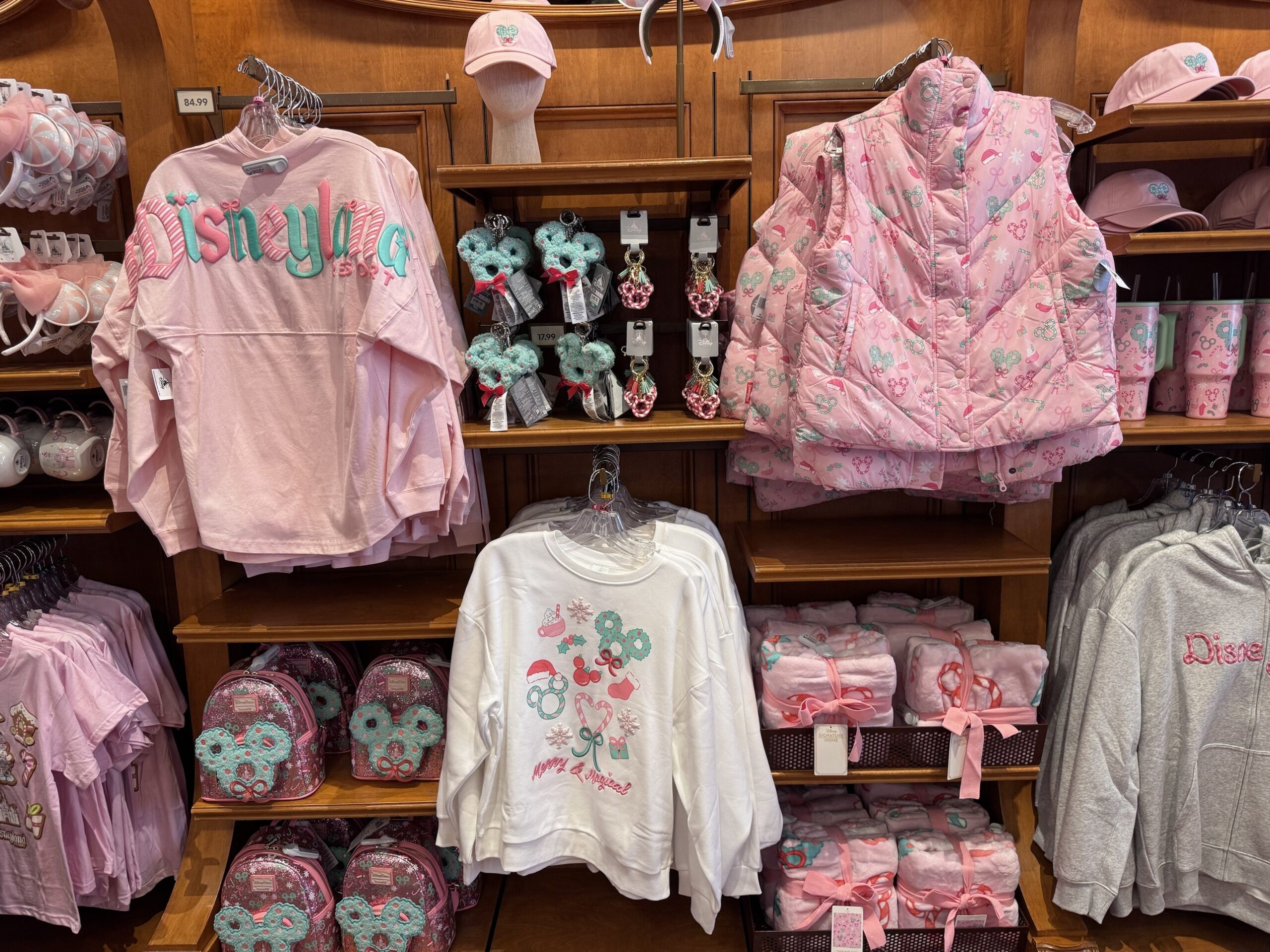 Pink and teal holiday merchandise wall at Disneyland with long-sleeve tops, bag charms, a puffer vest and Loungefly backpacks