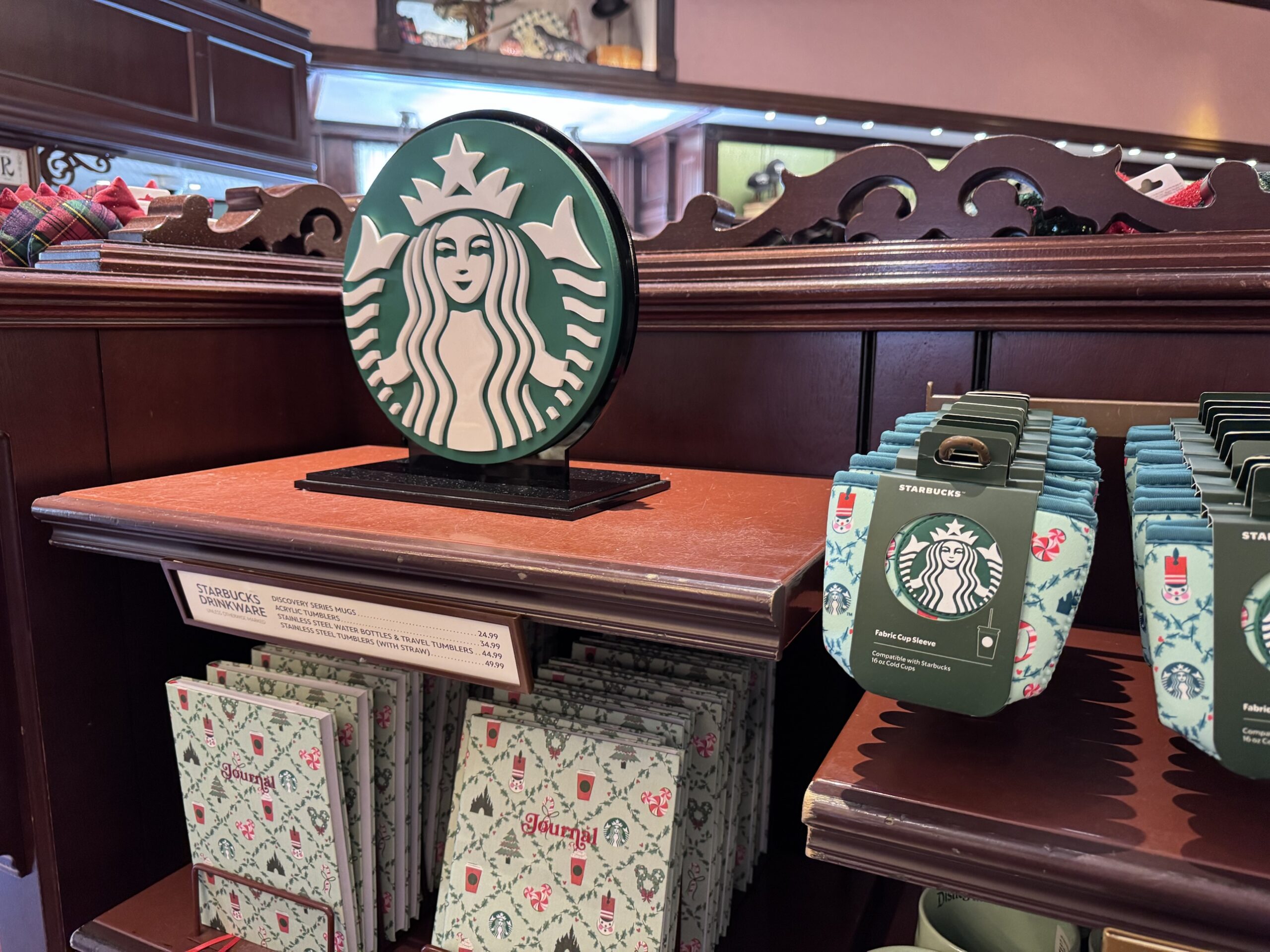 Starbucks drinkware price sign above holiday journals, with fabric cup sleeves on the shelf beside it at Disneyland