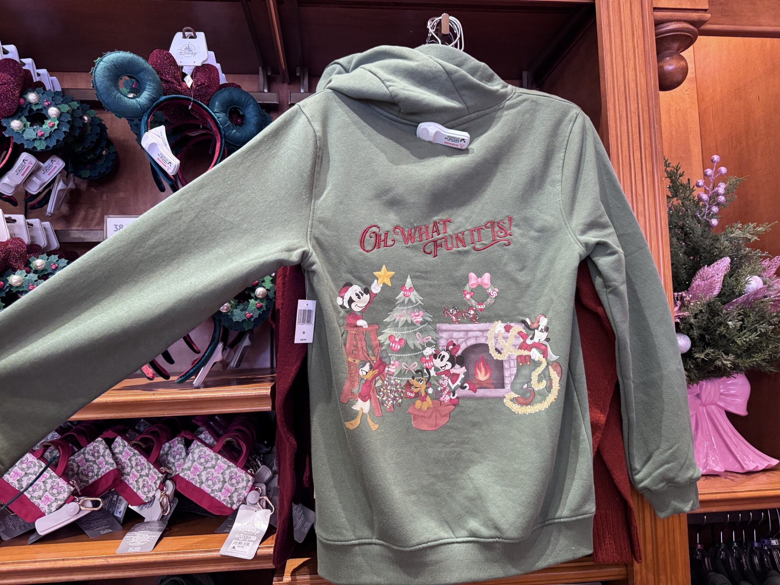 Back of a sage hoodie reading Oh What Fun It Is with Mickey, Minnie, Donald, Goofy and Pluto decorating a tree by a fireplace