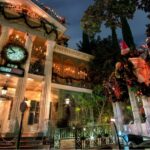The Haunted Mansion at Disneyland at dusk, decorated with dark garlands, jack-o'-lanterns and a striped bow, with a clock counting days to Christmas above the entrance